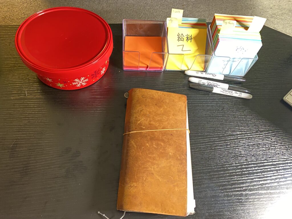 My desk, with a brown leather Travler's notebook, a red tupperware, and flashcard cubes full of kanji. The lighting is terrible.