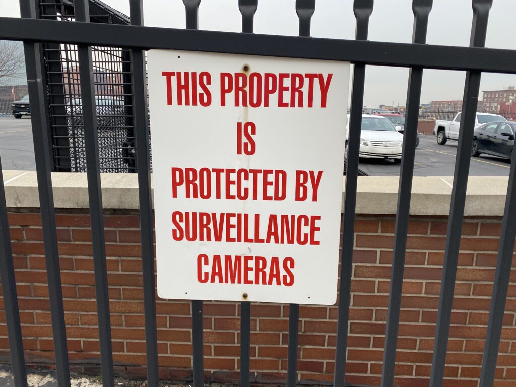 Red all-caps text on a sign that reads, "THIS PROPERTY IS PROTECTED BY SURVEILLANCE CAMERAS." It is hung from a metal fence around a parking garage.