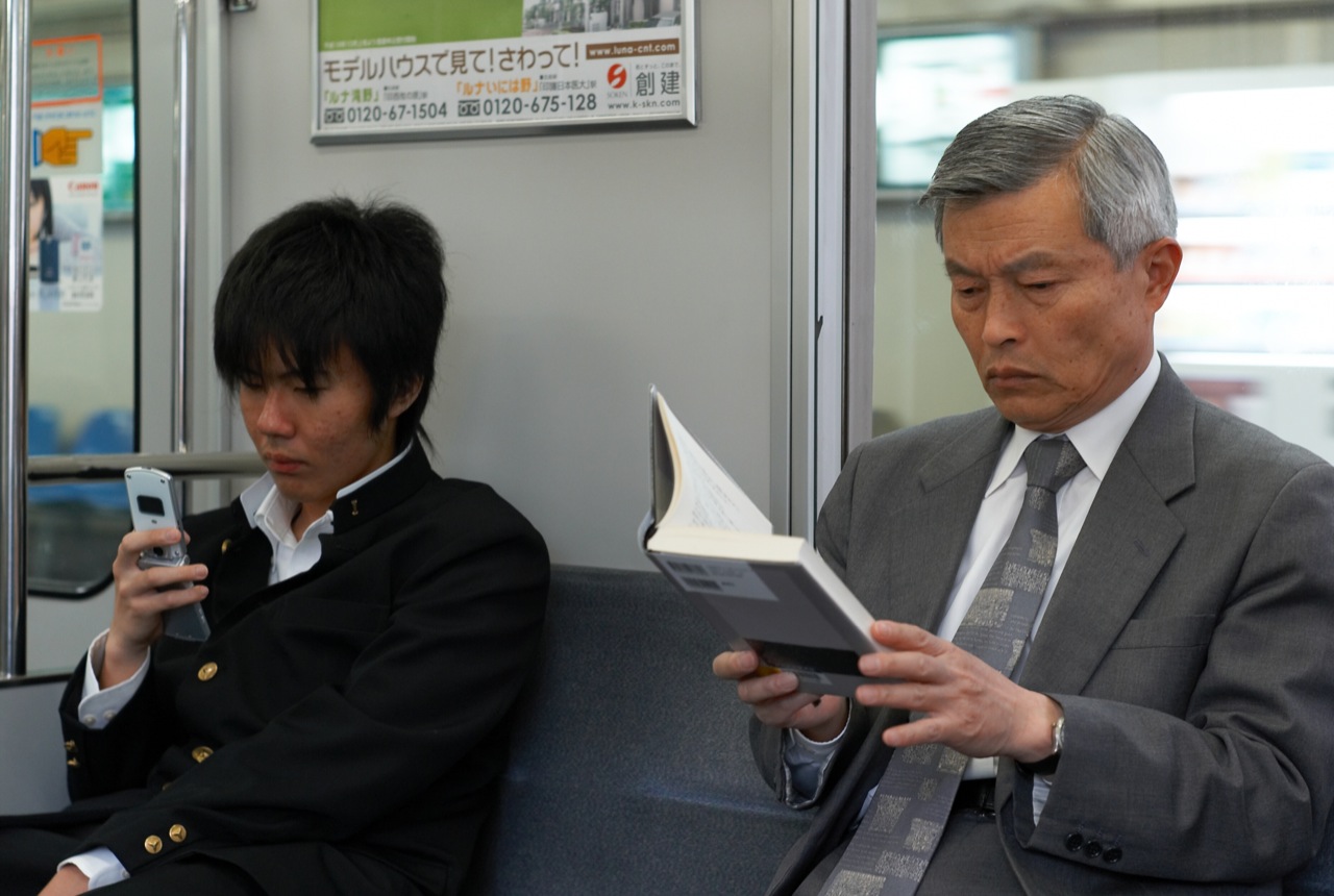 A young man on his phone next to an older man reading a book.